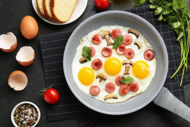 Photo of Tasty fried eggs with cut sausages and mushrooms in frying pan surrounded by ingredients on black table, flat lay