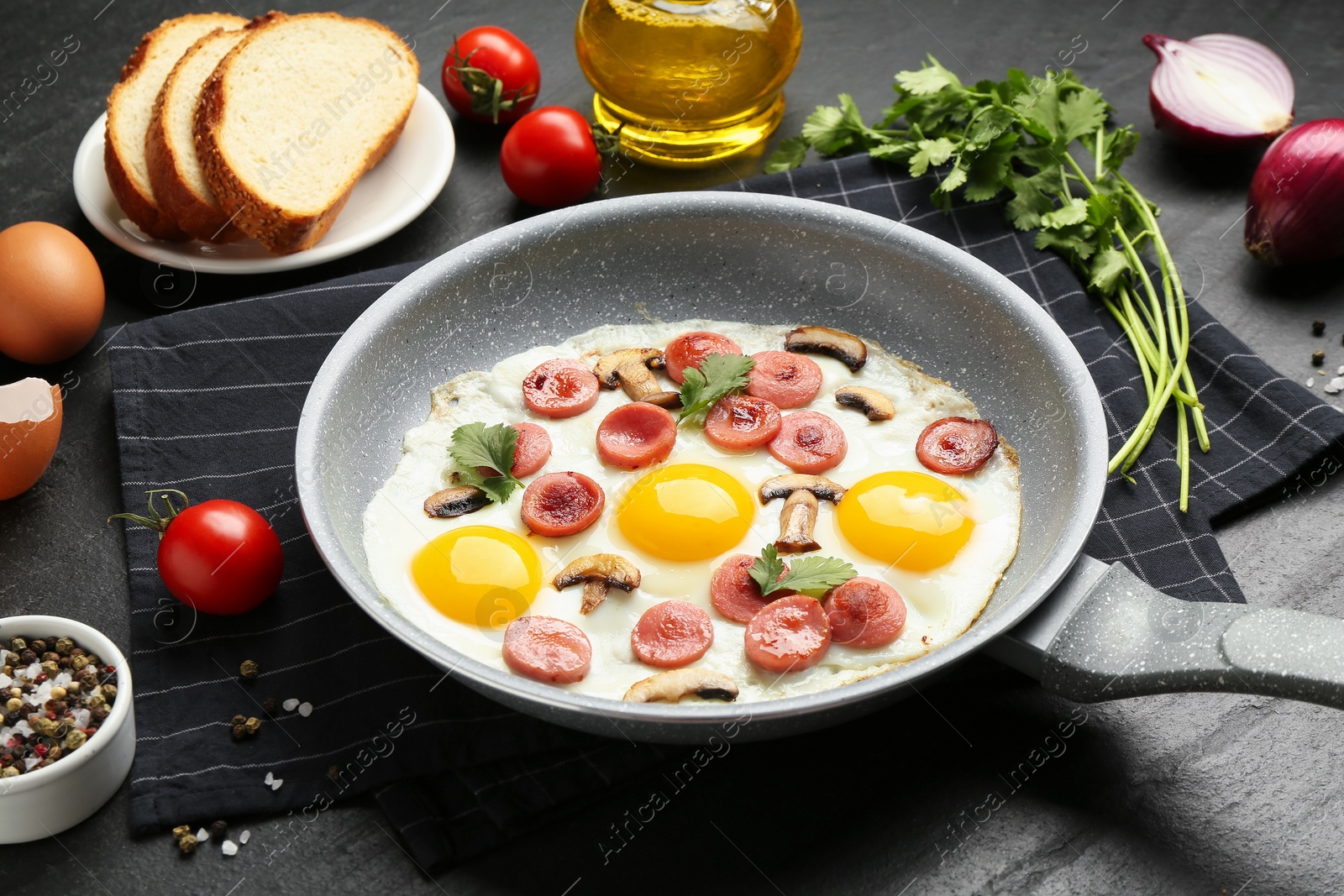 Photo of Tasty fried eggs with cut sausages and mushrooms in frying pan near ingredients on black table, closeup