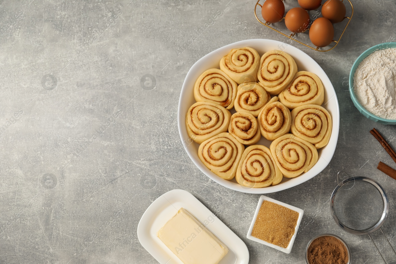 Photo of Raw cinnamon rolls in baking dish and different ingredients on grey table, flat lay. Space for text