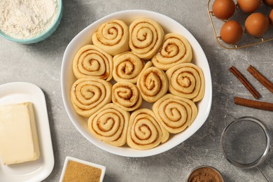 Photo of Raw cinnamon rolls in baking dish and different ingredients on grey table, flat lay