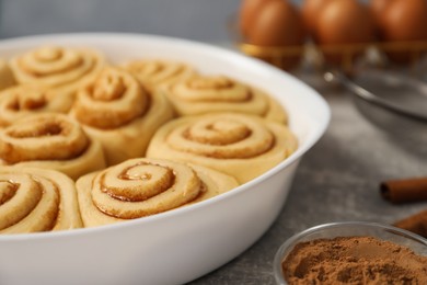 Photo of Raw cinnamon rolls in baking dish and cocoa powder on grey table, closeup