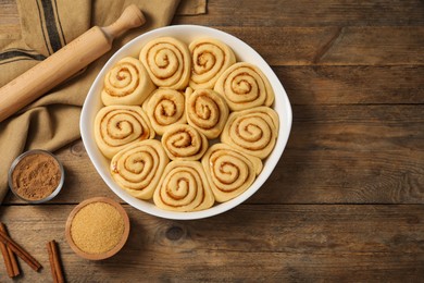 Raw cinnamon rolls in baking dish, rolling pin, cocoa powder and brown sugar on wooden table, flat lay. Space for text