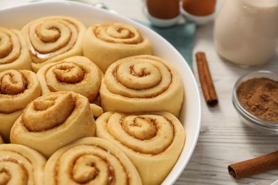 Photo of Raw cinnamon rolls in baking dish, sticks and cocoa powder on light wooden table, closeup