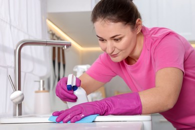 Photo of Woman using cleaning product while wiping sink with rag in kitchen