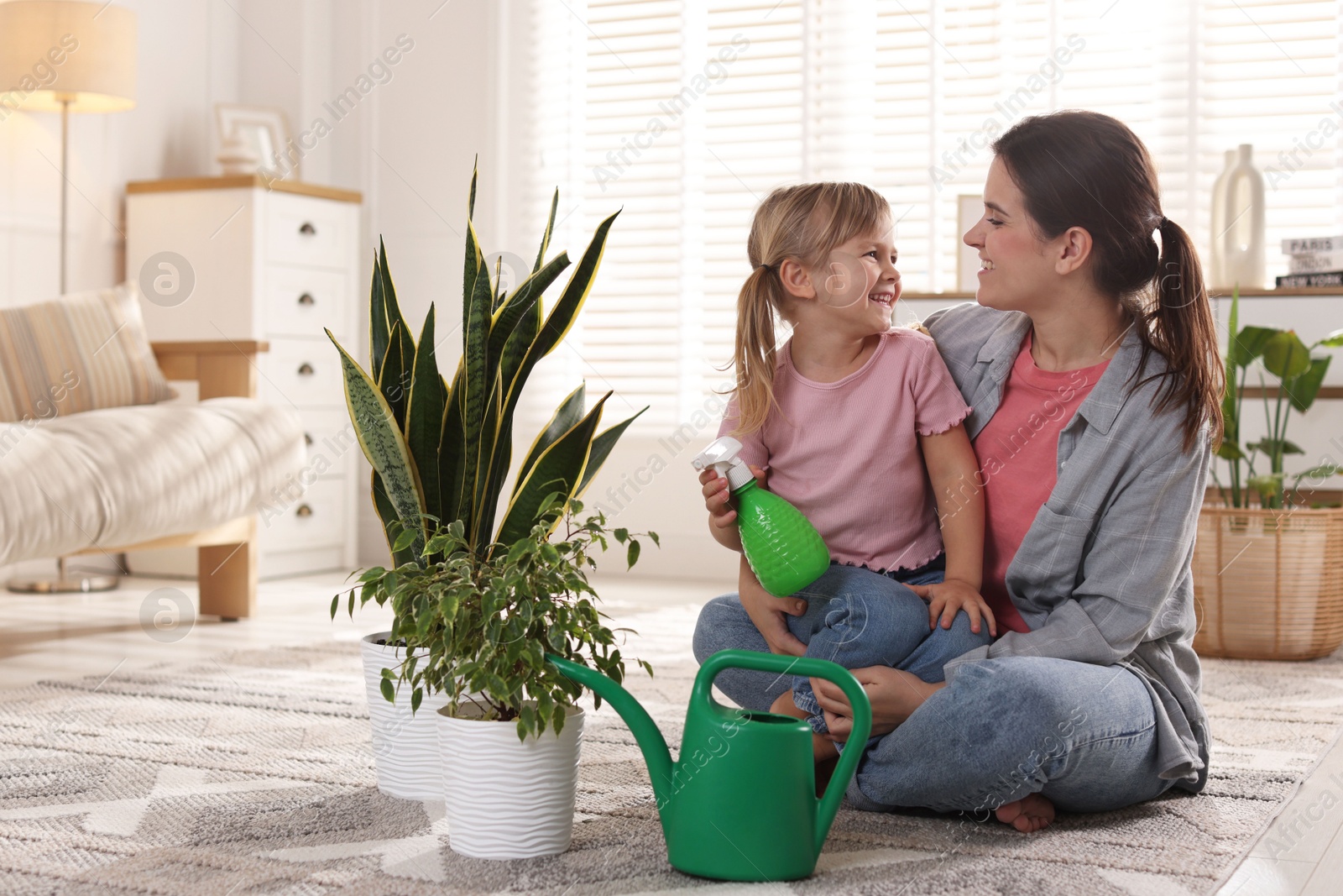 Photo of Little helper. Mother and her cute daughter with spray bottle near houseplants at home