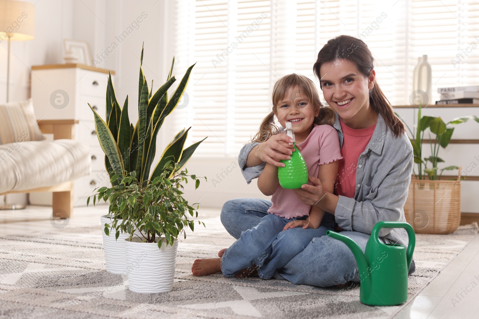 Photo of Little helper. Mother and her cute daughter with spray bottle near houseplants at home