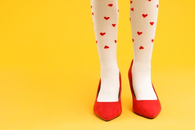 Photo of Little girl wearing oversized high heeled shoes on yellow background, closeup. Space for text