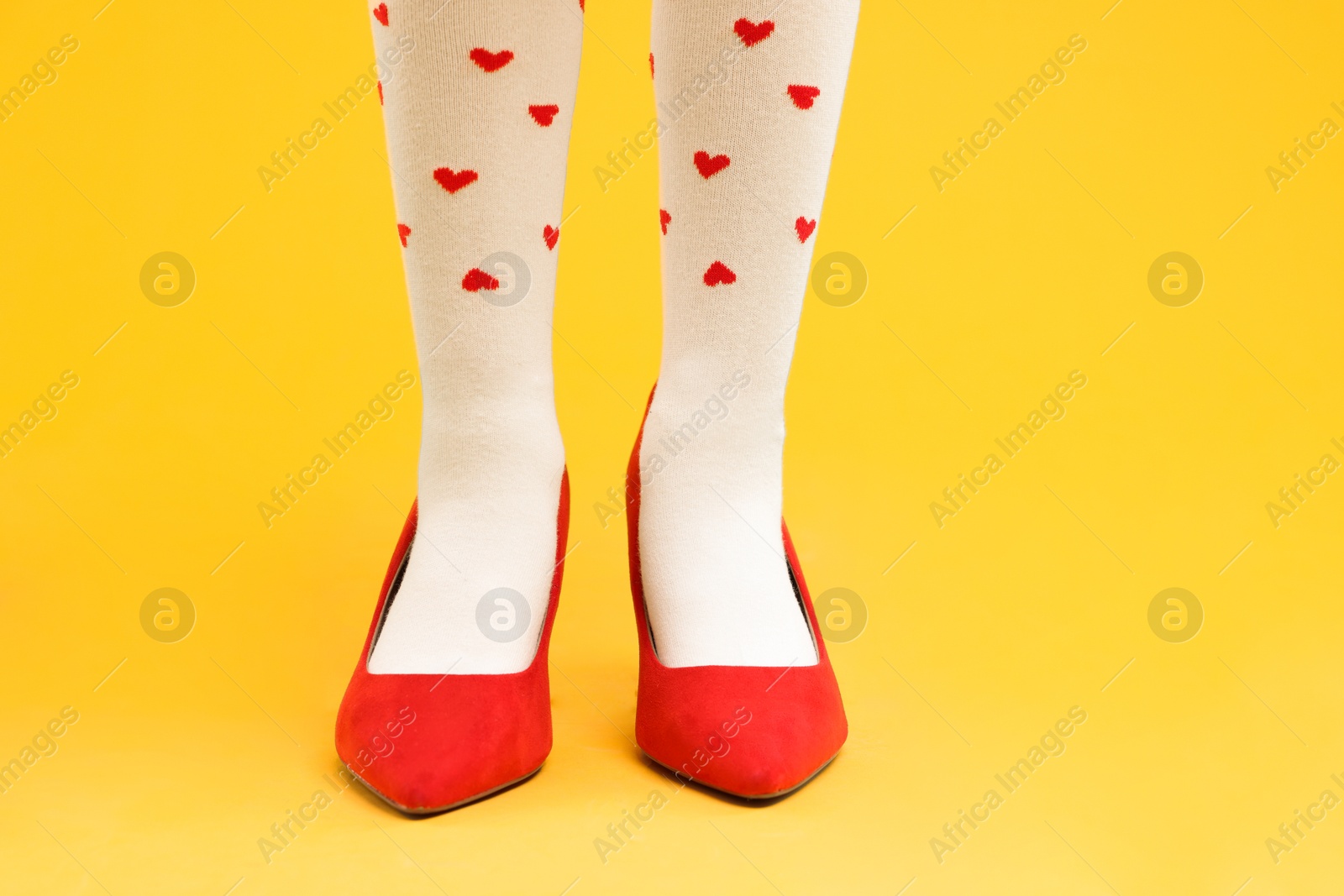 Photo of Little girl wearing oversized high heeled shoes on yellow background, closeup