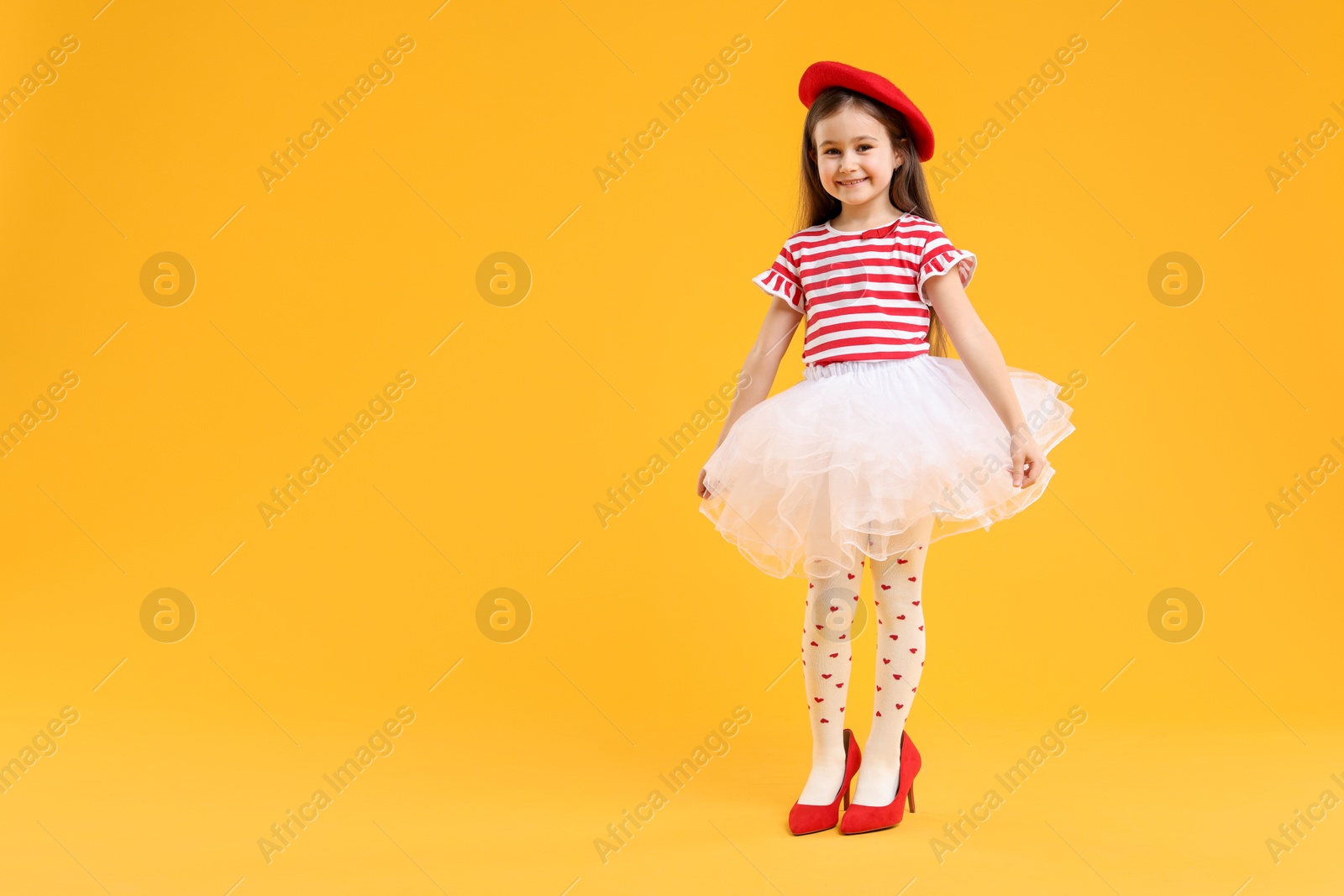 Photo of Stylish little girl wearing oversized high heeled shoes on orange background, space for text