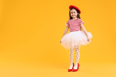 Photo of Stylish little girl wearing oversized high heeled shoes on orange background, space for text