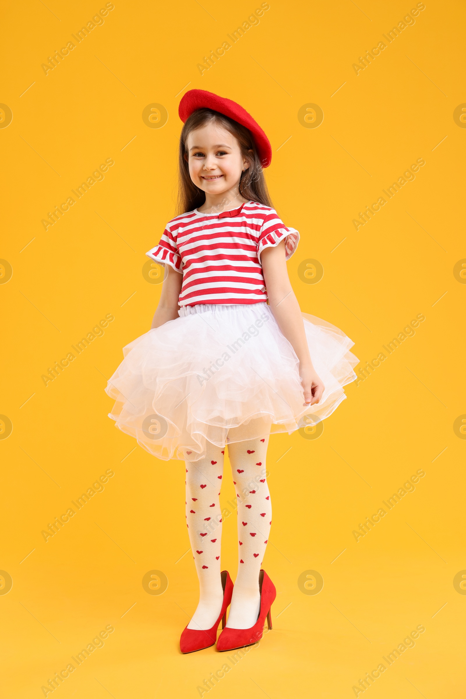 Photo of Stylish little girl wearing oversized high heeled shoes on orange background