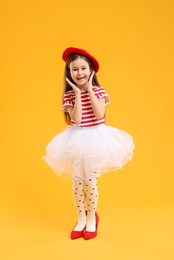 Photo of Stylish little girl wearing oversized high heeled shoes on orange background