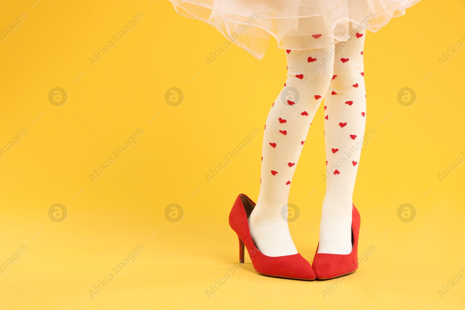 Photo of Little girl wearing oversized high heeled shoes on orange background, closeup. Space for text