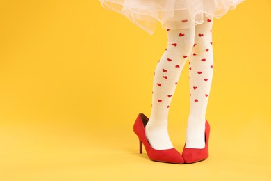 Photo of Little girl wearing oversized high heeled shoes on orange background, closeup. Space for text