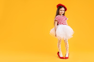 Photo of Stylish little girl wearing oversized high heeled shoes on orange background, space for text