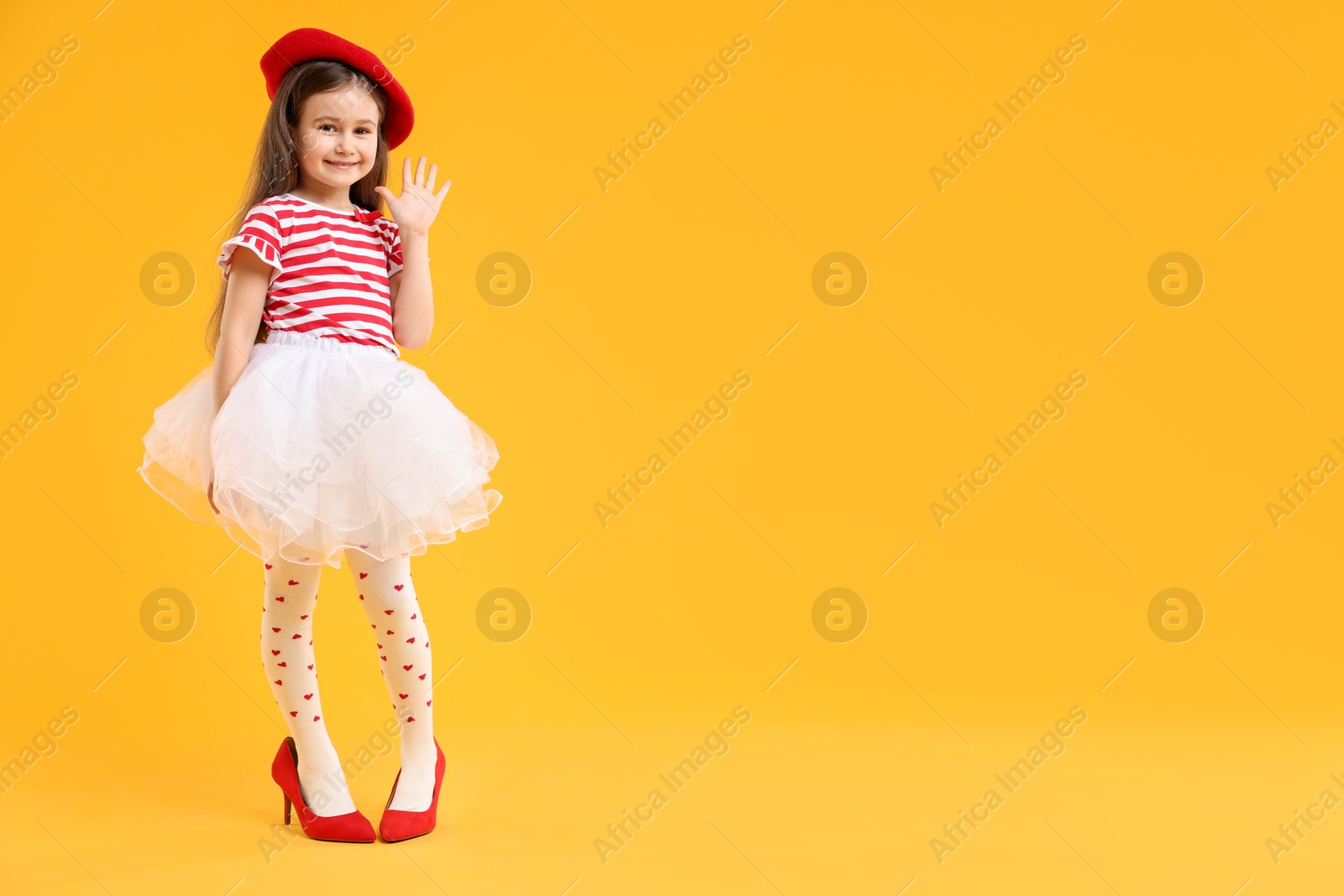 Photo of Stylish little girl wearing oversized high heeled shoes on orange background, space for text