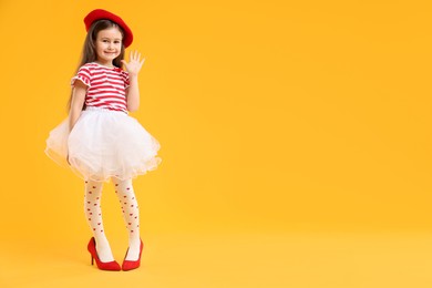 Photo of Stylish little girl wearing oversized high heeled shoes on orange background, space for text