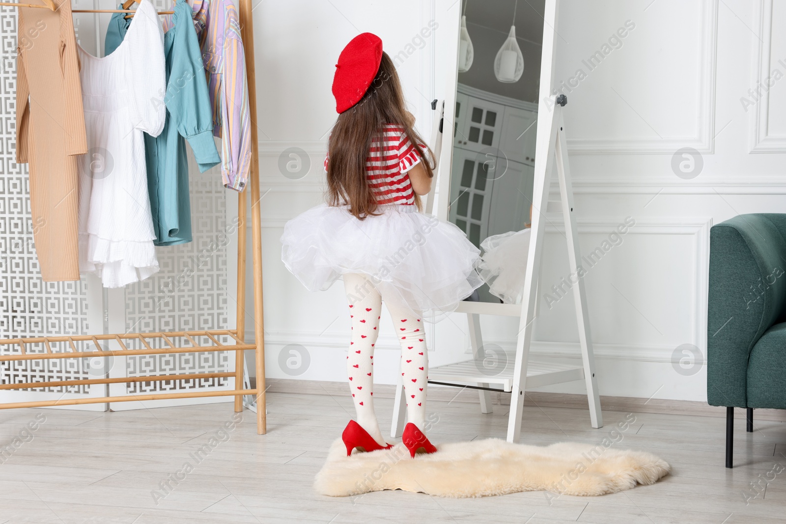 Photo of Stylish little girl wearing oversized high heeled shoes near mirror indoors