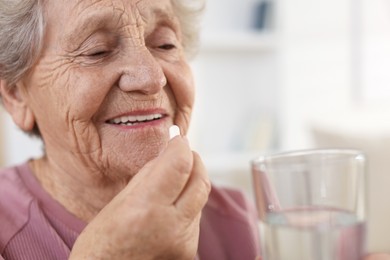 Smiling senior woman with glass of water taking pill at home, closeup