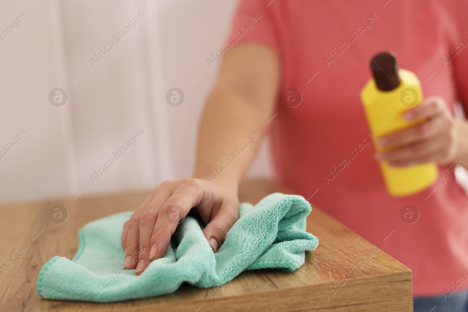 Photo of Woman holding bottle of cleaning product while polishing wooden table with rag at home, closeup