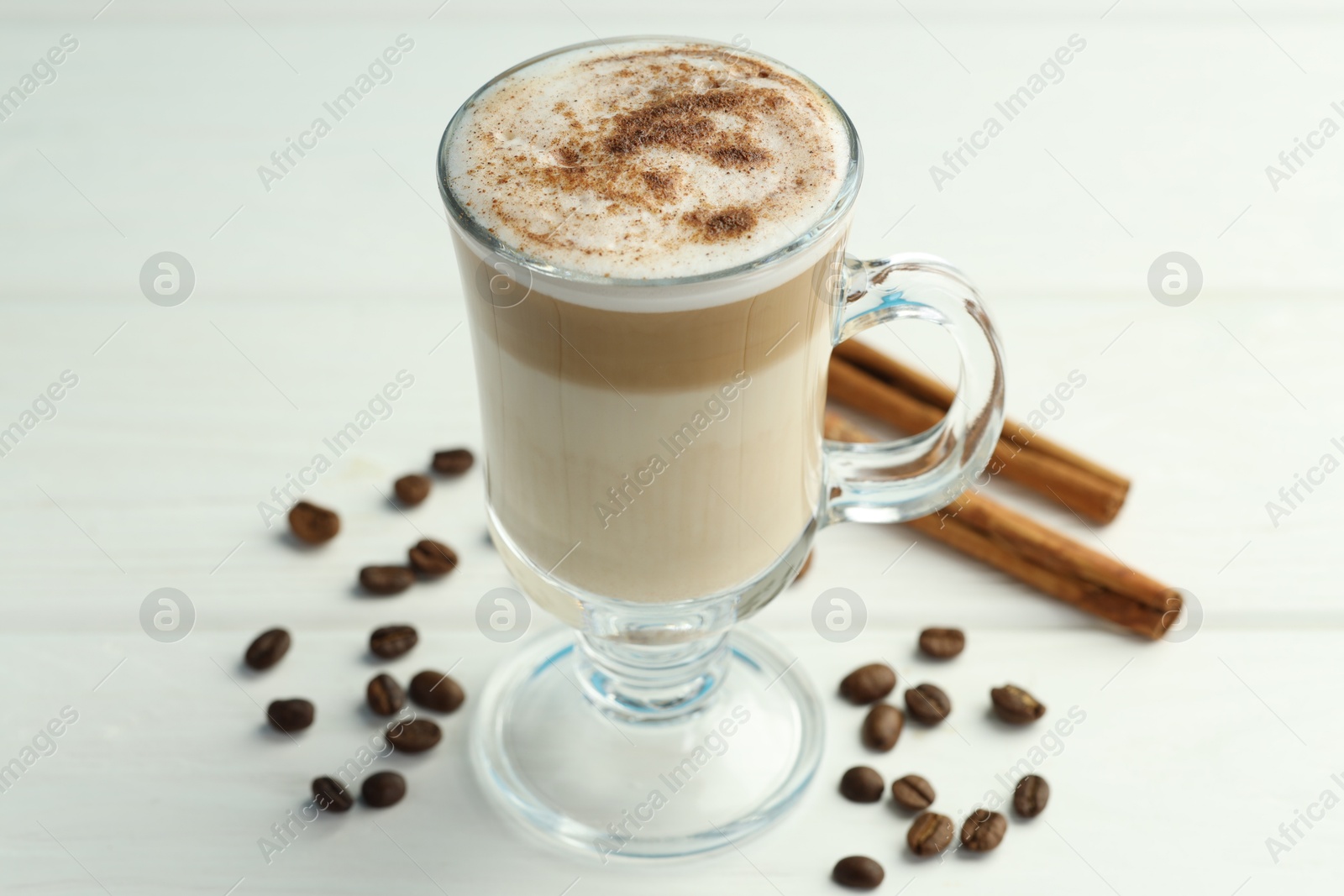 Photo of Tasty latte macchiato in glass cup, coffee beans and cinnamon on white wooden table, closeup