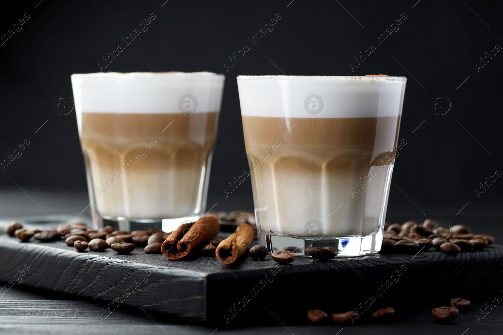 Photo of Tasty latte macchiato in glasses, coffee beans and cinnamon on black wooden table, closeup