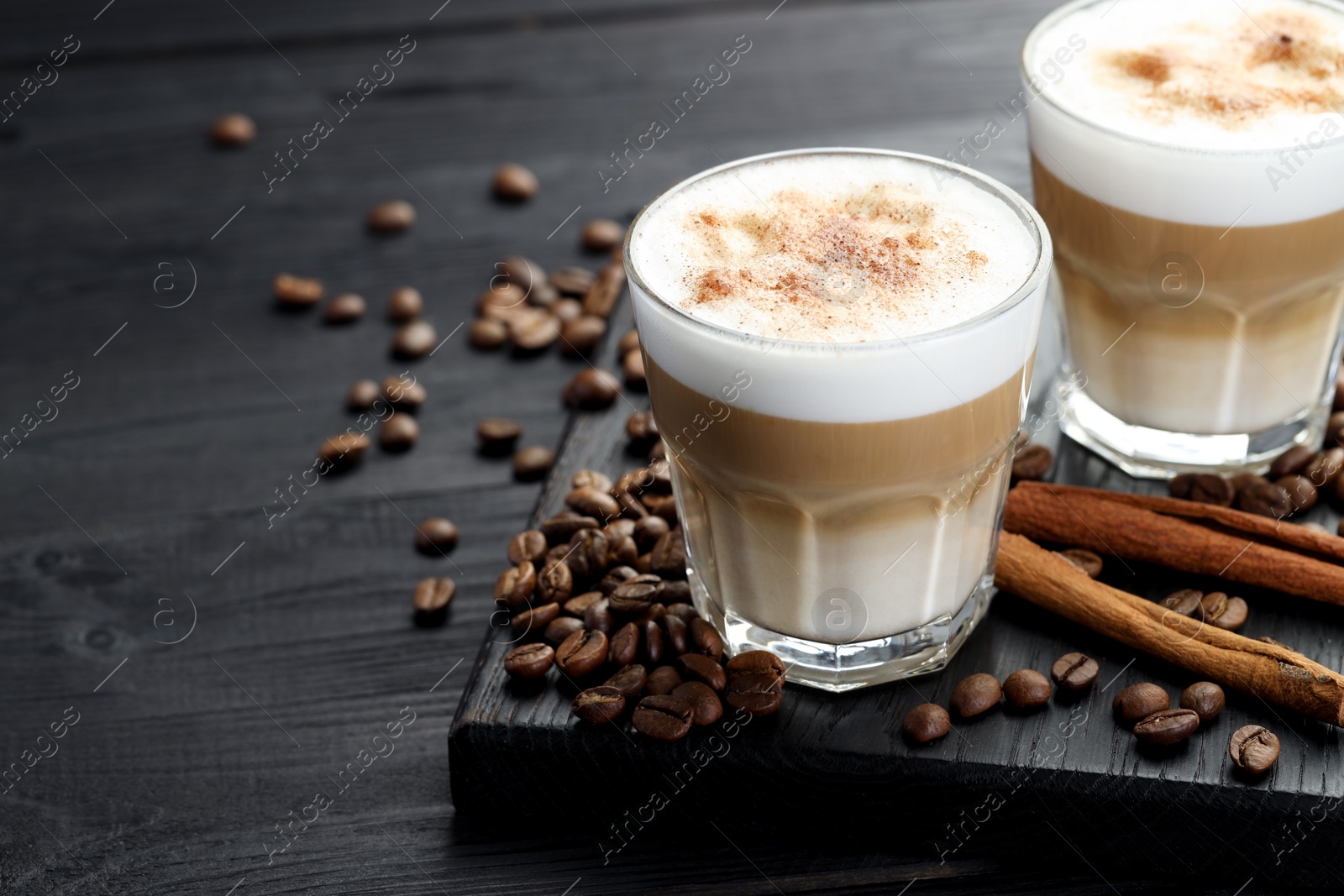 Photo of Tasty latte macchiato in glasses, coffee beans and cinnamon on black wooden table, closeup. Space for text