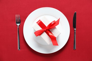 Gift and setting for romantic dinner on red background, top view