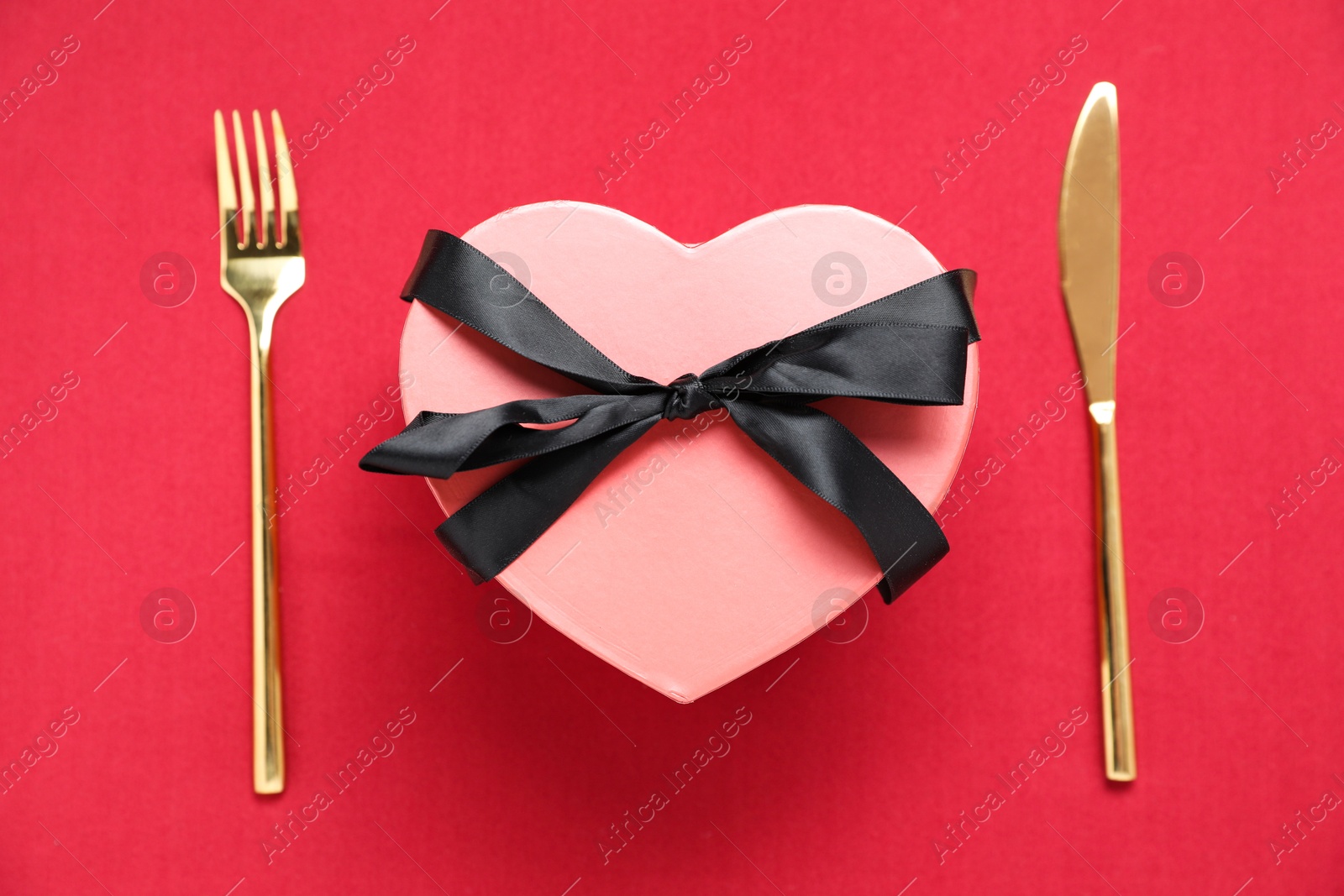 Photo of Heart shaped romantic gift and cutlery on red background, top view