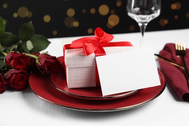 Gift box, plates, roses and blank card on white table against black background, closeup. Table setting for romantic dinner