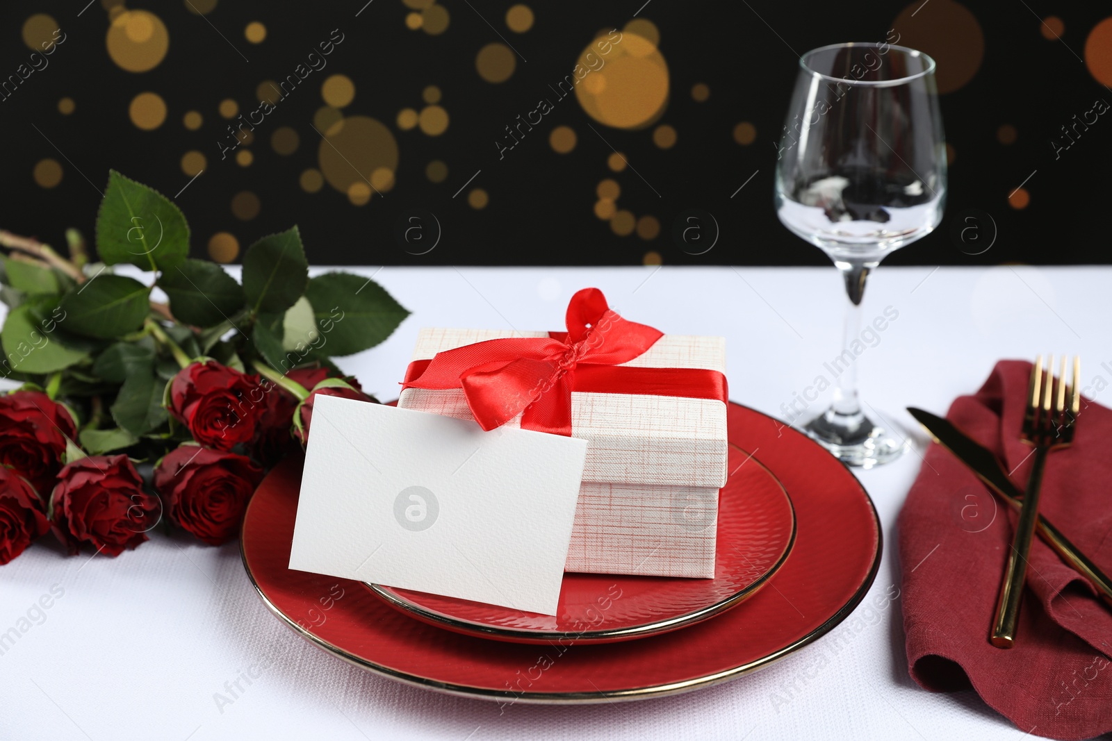 Photo of Gift box, plates, roses and blank card on white table against black background. Table setting for romantic dinner