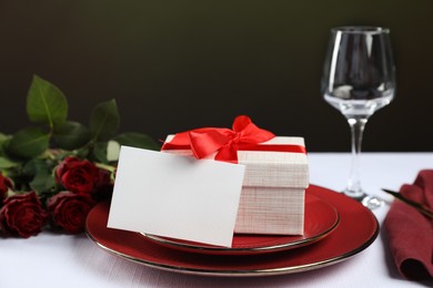 Gift box, plates, roses and blank card on white table against black background, closeup. Table setting for romantic dinner