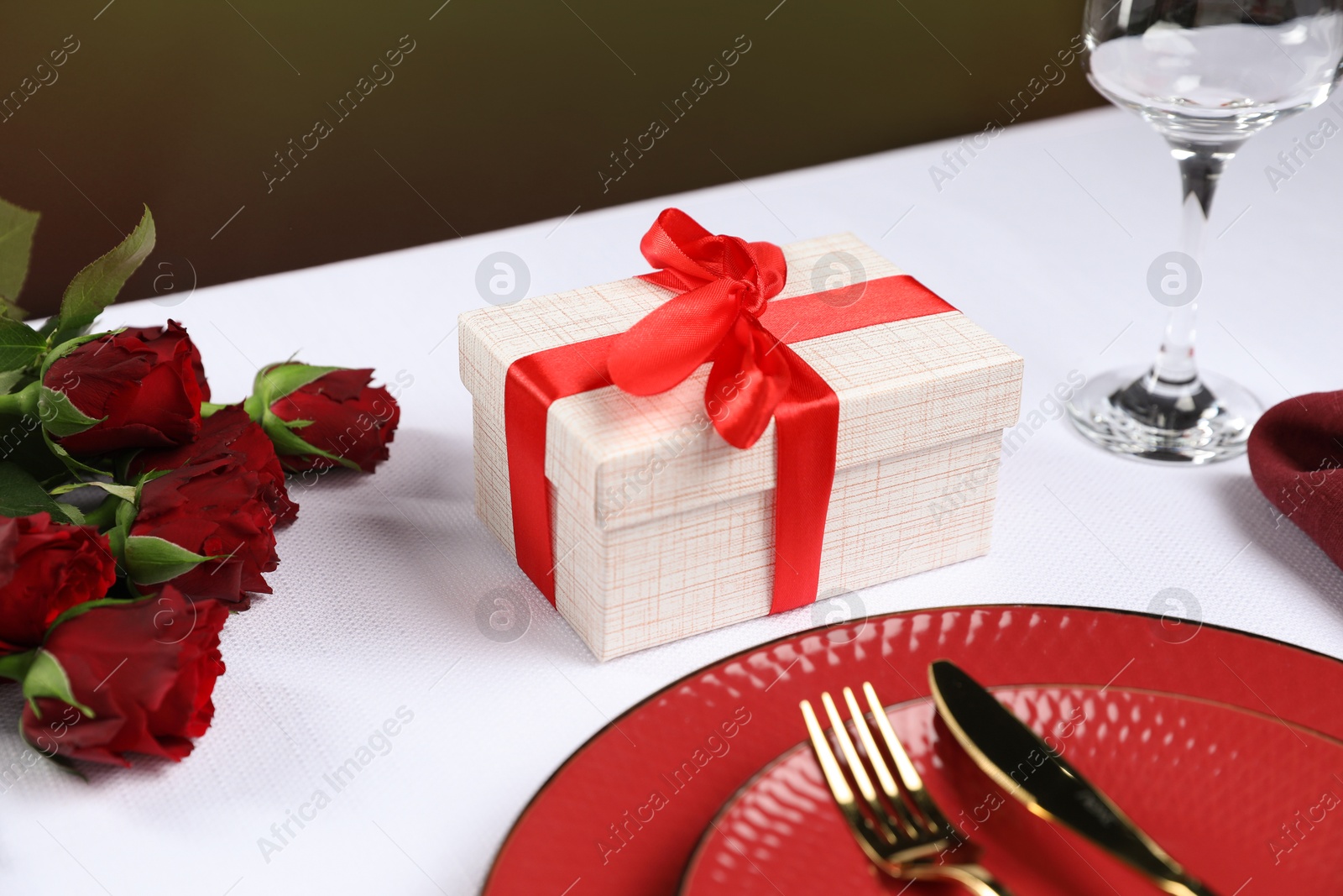 Photo of Gift box, plates, cutlery and roses on white table against black background, closeup. Table setting for romantic dinner