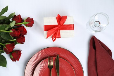 Gift box, plates, cutlery and roses on white table, flat lay. Beautiful table setting for romantic dinner