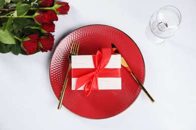 Photo of Gift box, plate, cutlery and roses on white table, flat lay. Beautiful table setting for romantic dinner