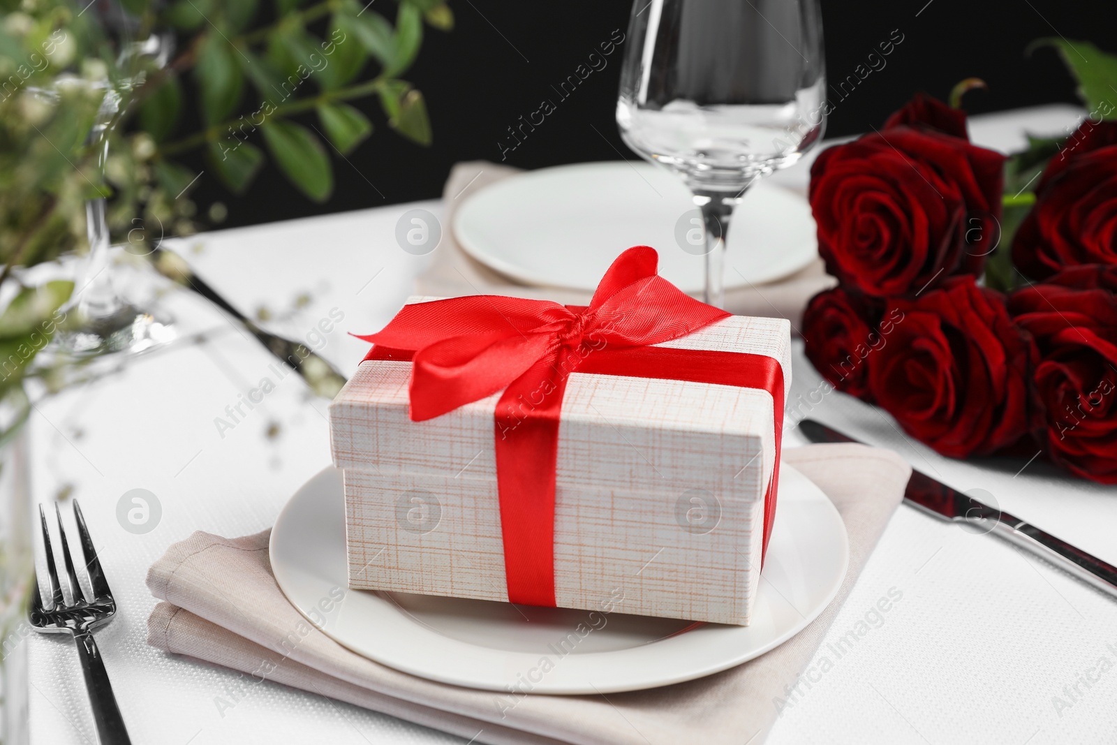 Photo of Gift box, plates, cutlery and roses on white table, closeup. Beautiful table setting for romantic dinner