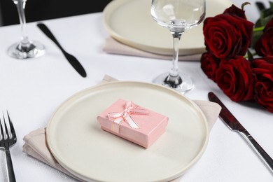 Photo of Gift box, plates, cutlery and roses on white table, closeup. Beautiful table setting for romantic dinner