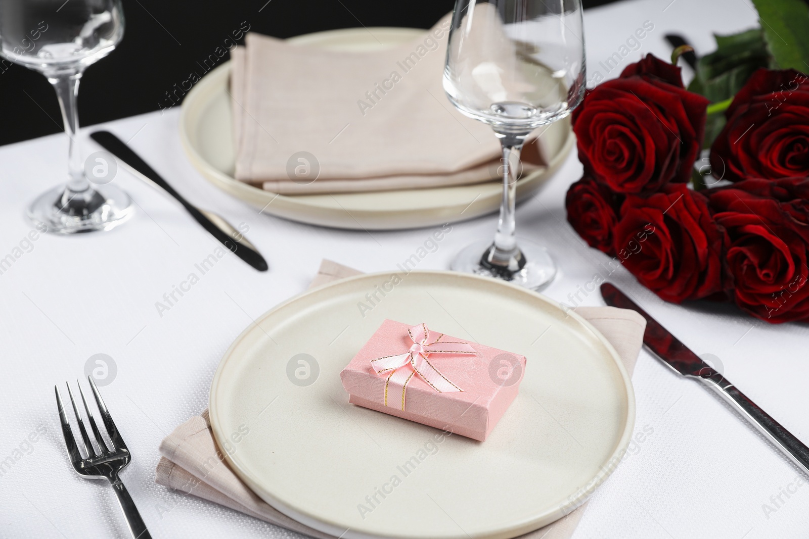 Photo of Gift box, plates, cutlery and roses on white table, closeup. Beautiful table setting for romantic dinner