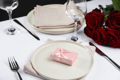 Photo of Gift box, plates, cutlery and roses on white table, closeup. Beautiful table setting for romantic dinner