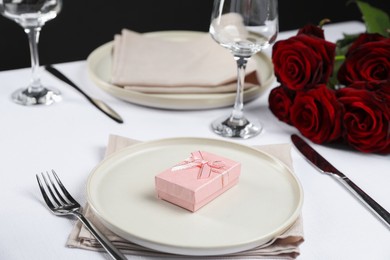 Gift box, plates, cutlery and roses on white table, closeup. Beautiful table setting for romantic dinner