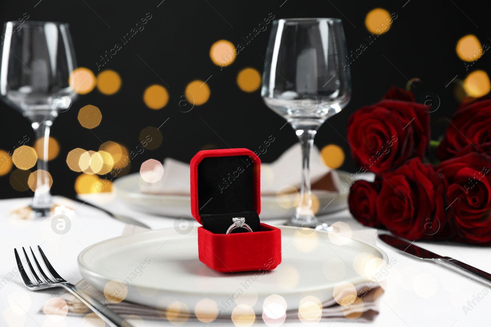 Photo of Engagement ring in box and tableware on white tablecloth, closeup. Table setting for romantic dinner