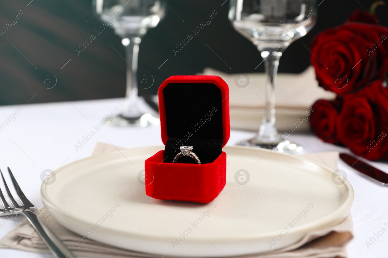 Photo of Engagement ring in box and tableware on white tablecloth, closeup. Table setting for romantic dinner