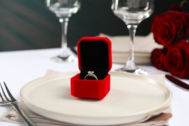 Engagement ring in box and tableware on white tablecloth, closeup. Table setting for romantic dinner