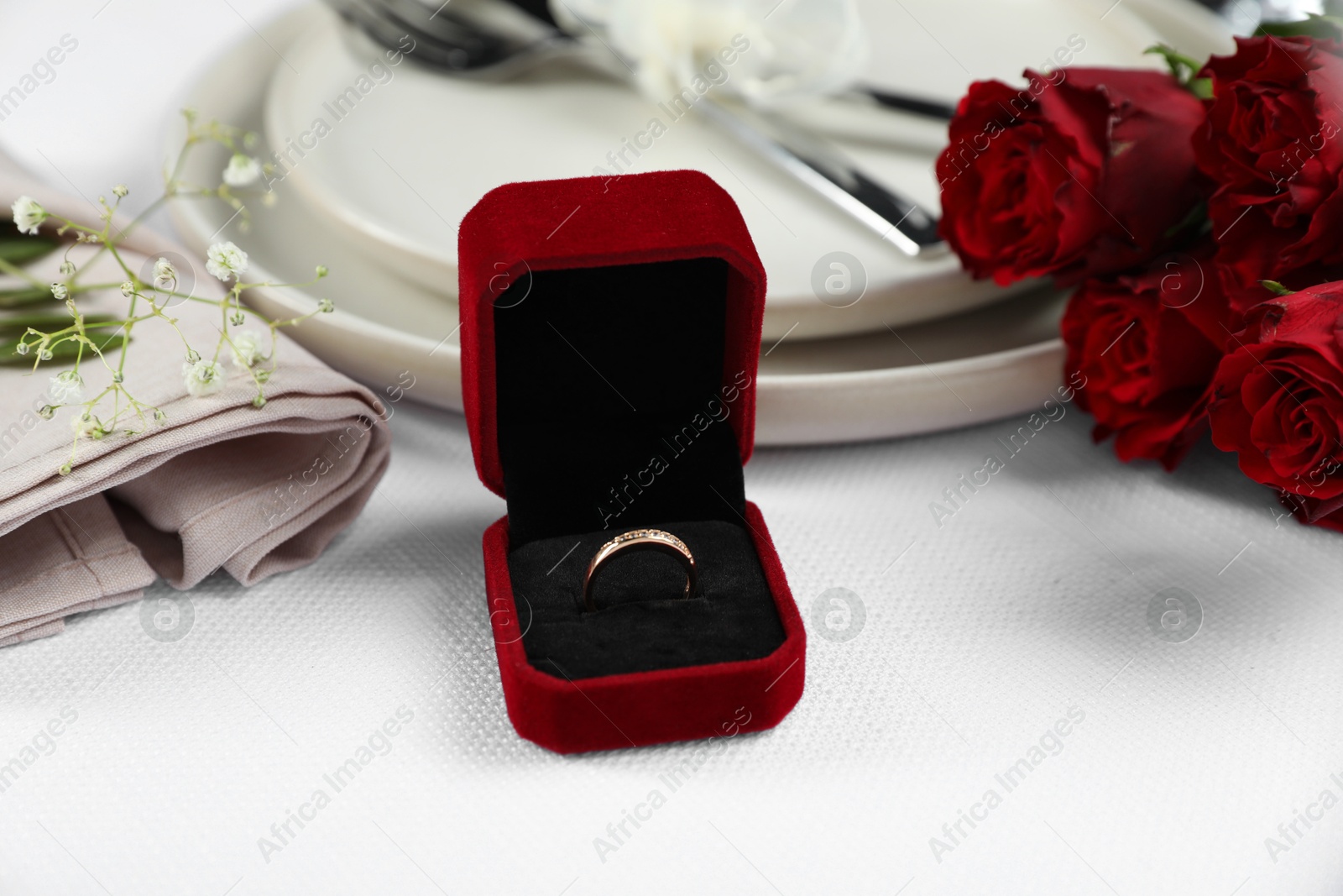 Photo of Engagement ring in box, rose flowers and tableware on white tablecloth, closeup. Romantic dinner