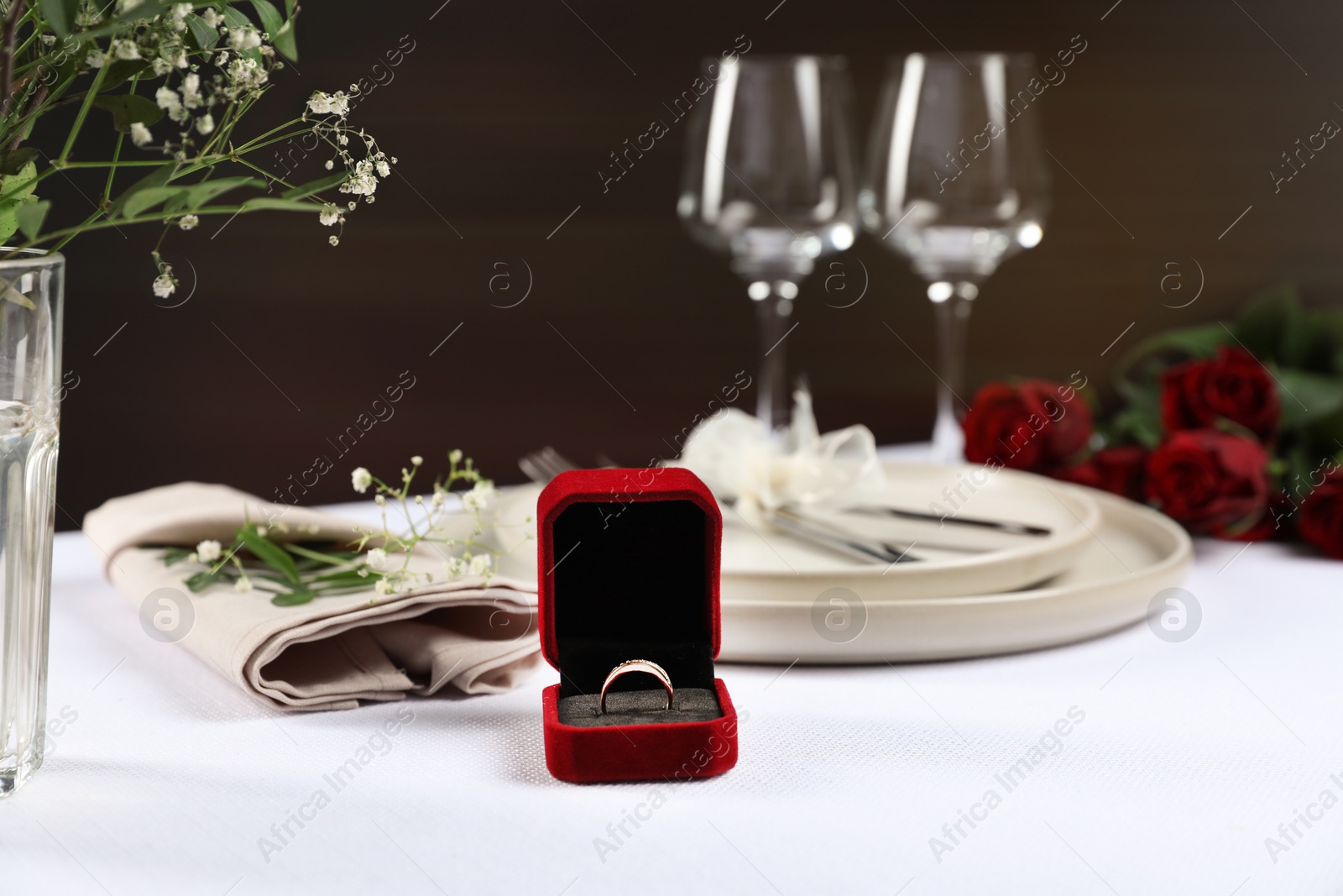 Photo of Engagement ring in box, flowers and tableware on white tablecloth. Romantic dinner