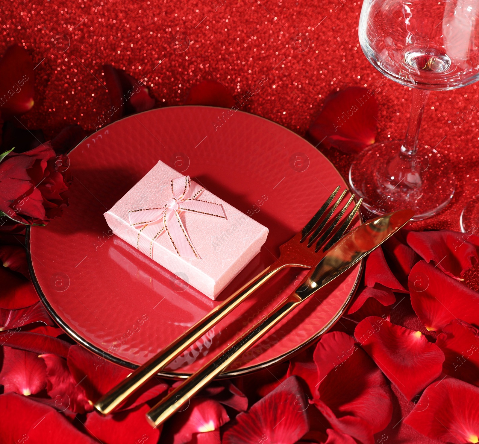 Photo of Gift box, plate, cutlery and roses on red glitter background. Beautiful table setting for romantic dinner