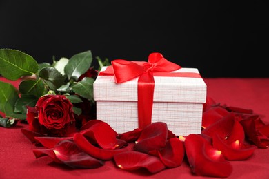 Beautiful gift box among rose flowers and petals on red table against black background. Romantic present