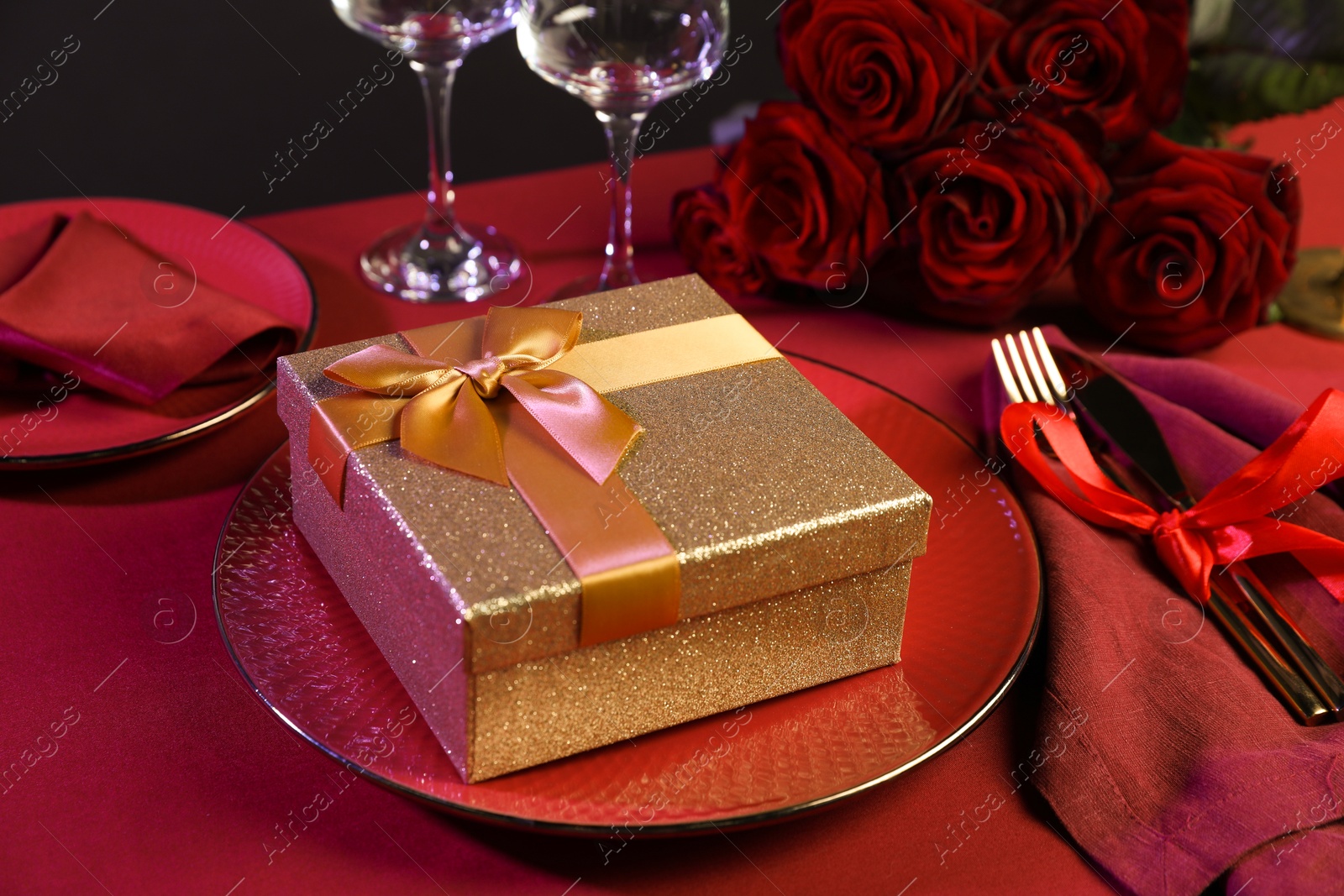 Photo of Gift box, tableware and roses on red table, closeup. Table setting for romantic dinner