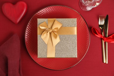 Gift box and tableware on red table, flat lay. Table setting for romantic Valentine's day celebration