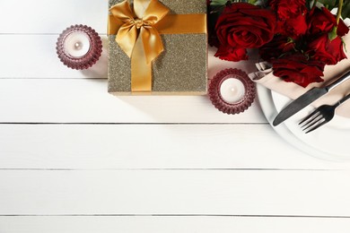 Beautiful table setting with romantic gift, flowers and burning candles on white wooden background, flat lay. Space for text